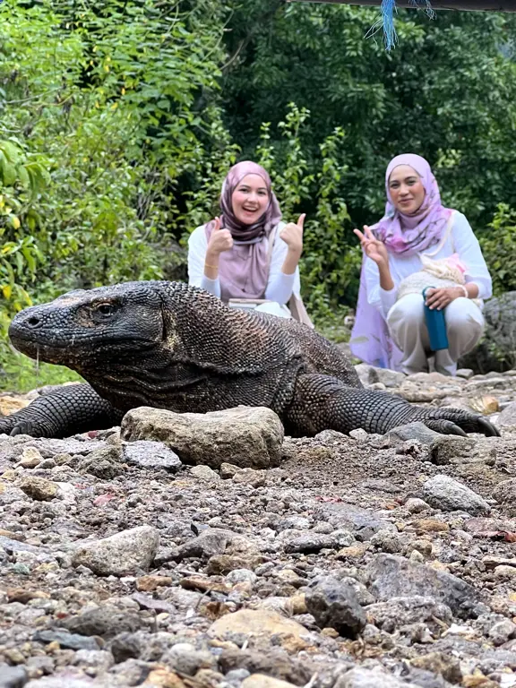 Dua tamu berfoto di belakang komodo di tepi hutan