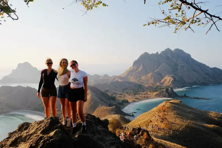 Tiga tamu tersenyum di batu puncak Padar dengan pantai melengkung di bawah