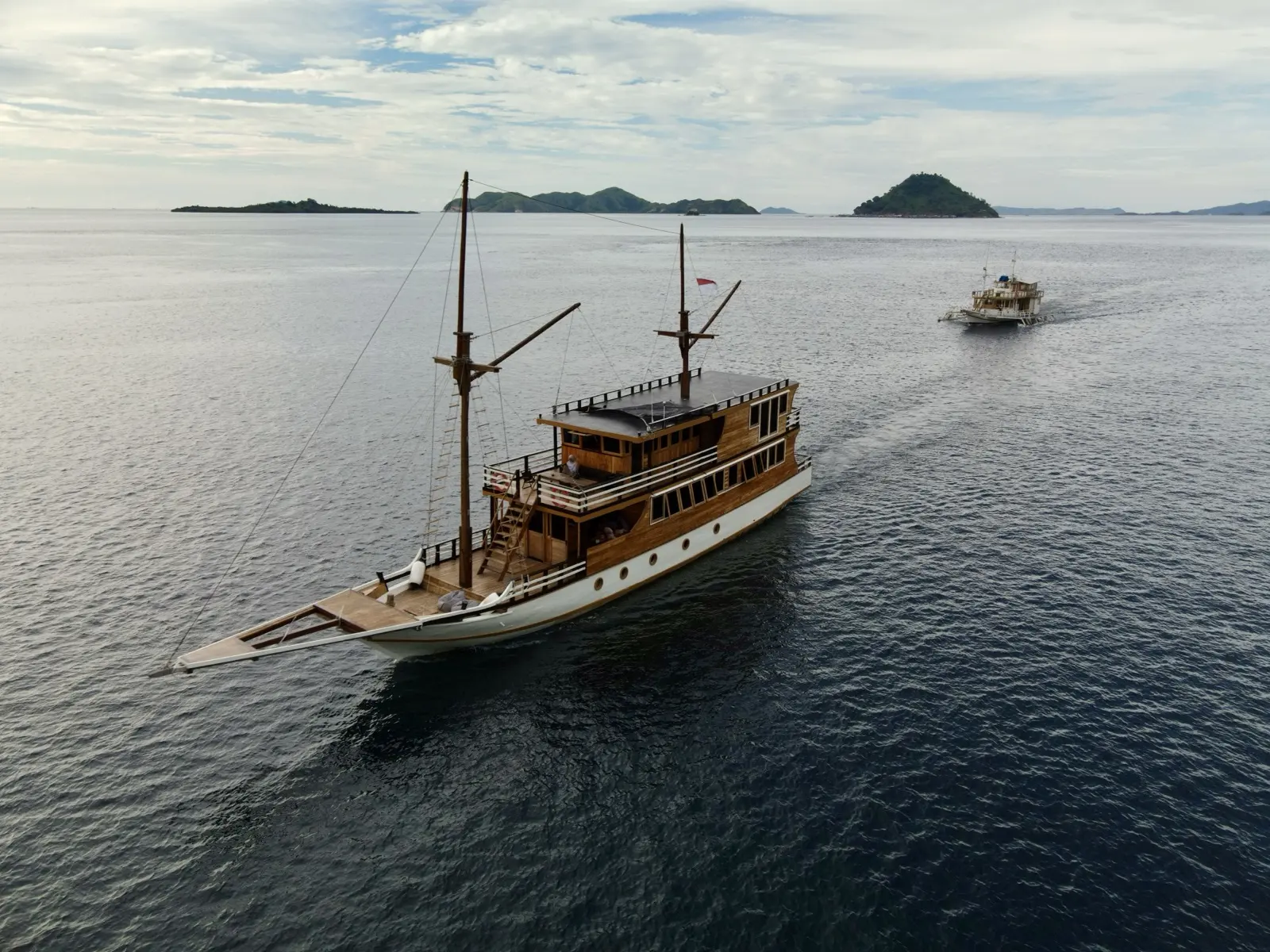 Kapal phinisi kayu Bajo Sunset Voyages dari udara, dua tiang dan bendera Merah Putih, berlayar di depan bukit Taman Nasional Komodo