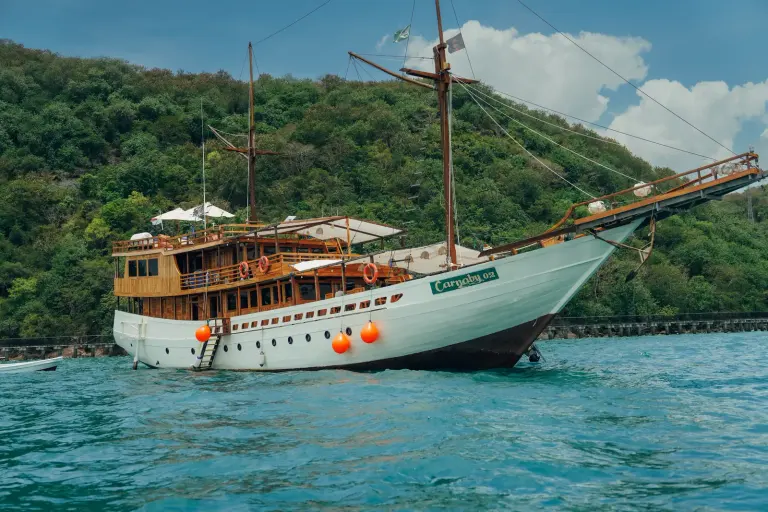 The white-hulled two-masted Carnaby II phinisi at anchor in a green Labuan Bajo bay