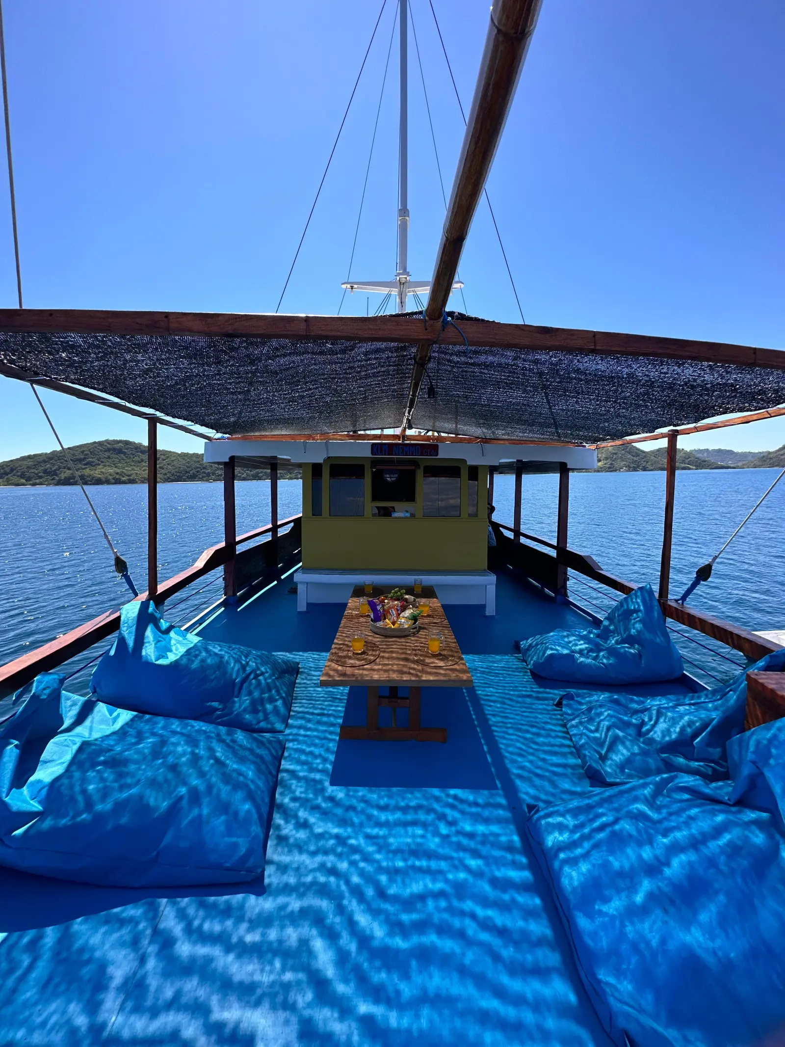 Ruang santai dek atas Nemmo dengan beanbag biru dan meja rendah, tiang layar dan laut di belakang