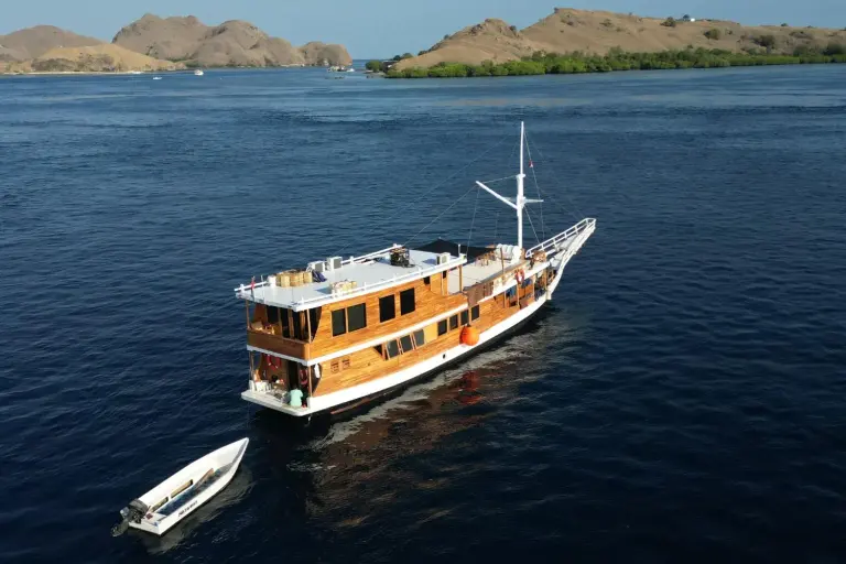 Foto udara phinisi kayu Segara di perairan biru Komodo, tender di sampingnya dan bukit cokelat di latar
