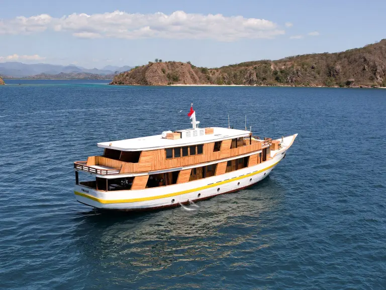 Aerial view of the wooden boat Vhalé, white hull with a yellow stripe, sailing near the hills of Komodo National Park